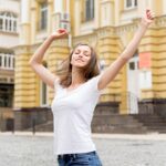 Femme en ville, qui écarte les bras avec les yeux fermés, et qui semble apaisée, équilibrée.