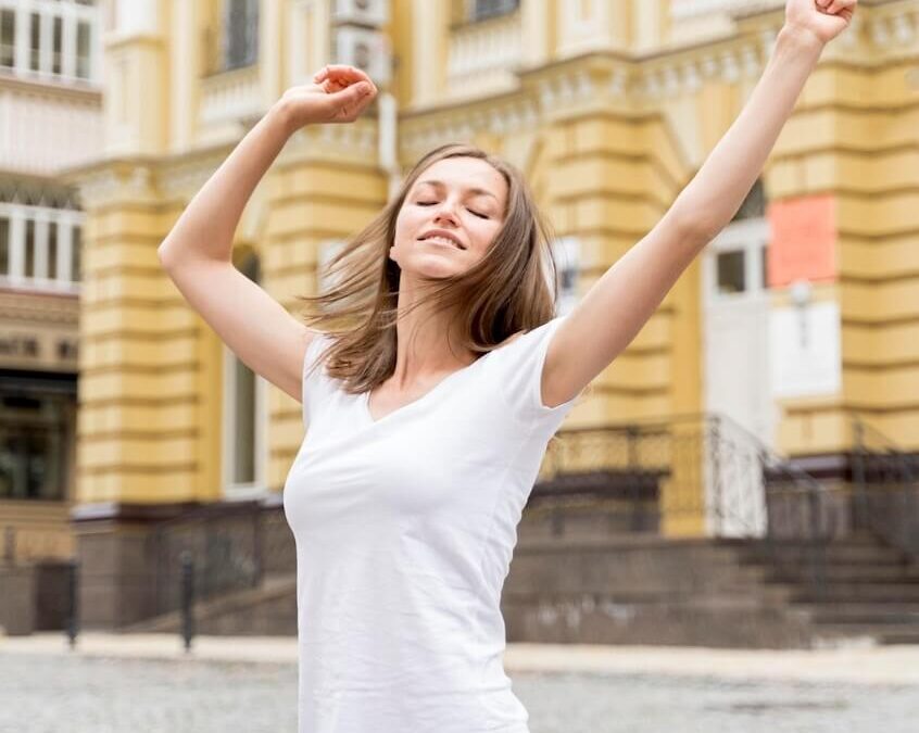 Femme en ville, qui écarte les bras avec les yeux fermés, et qui semble apaisée, équilibrée.