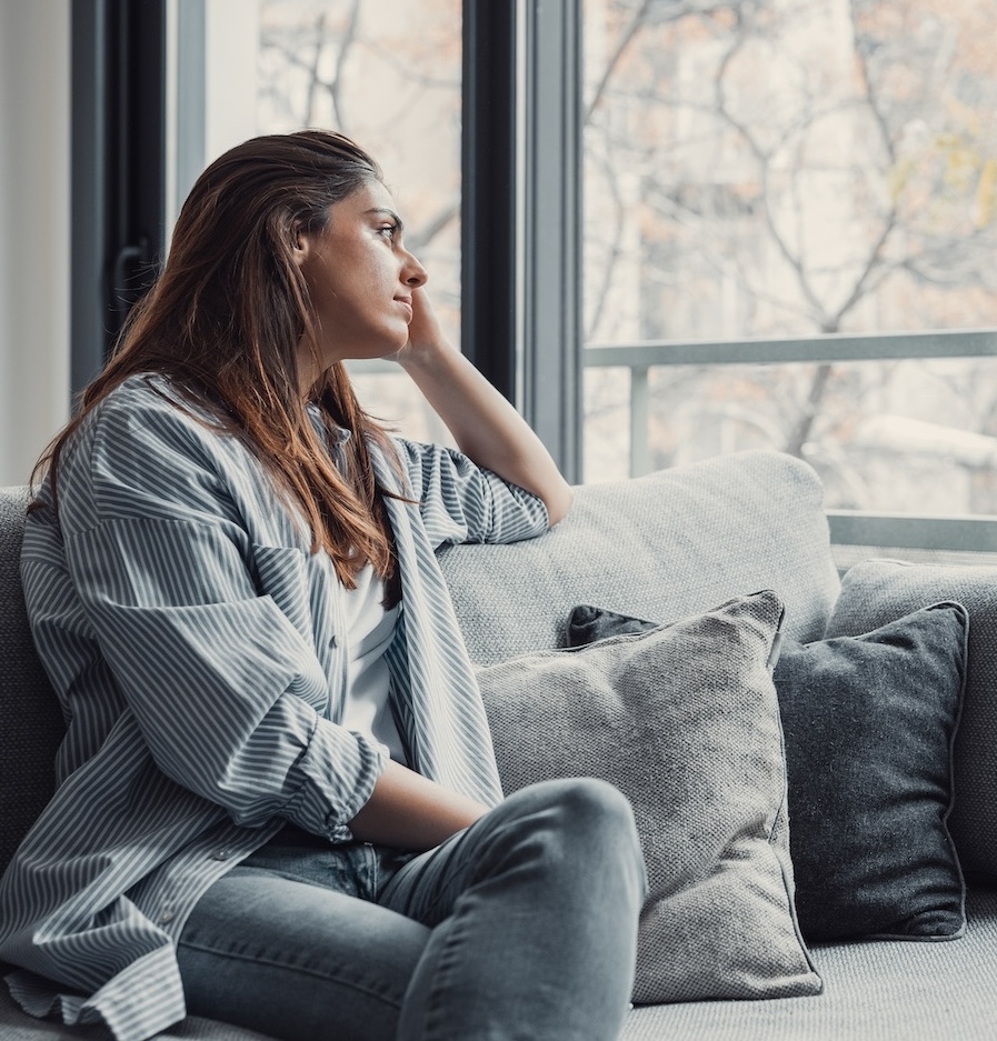 Femme assise sur son canapé qui regarde par la fenêtre, qui est anxieuse et triste.