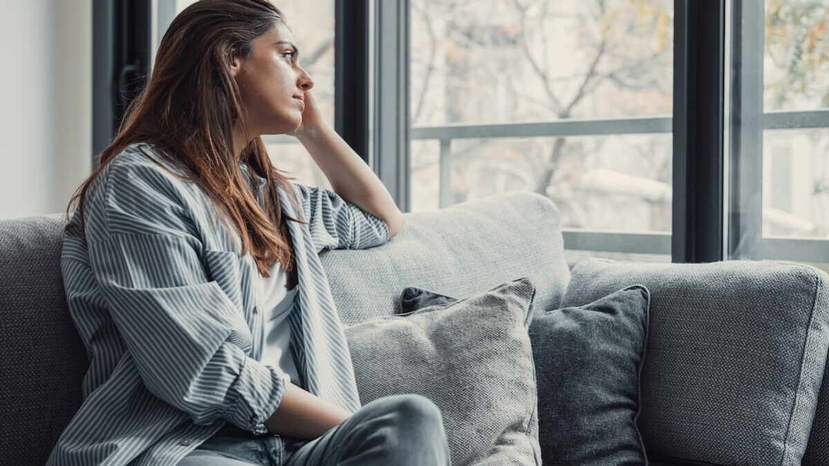 Femme qui a des pensées négatives, assise sur son canapé en regardant par la fenêtre