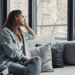 Femme qui a des pensées négatives, assise sur son canapé en regardant par la fenêtre